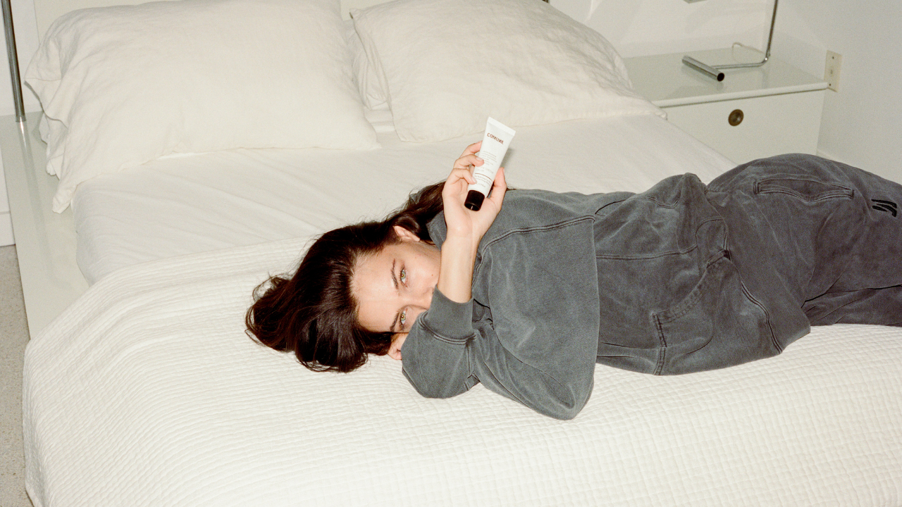 Comune model wearing COMUNE clothing lying on a bed showing 'COMUNE" Hydra Shroom Cream.