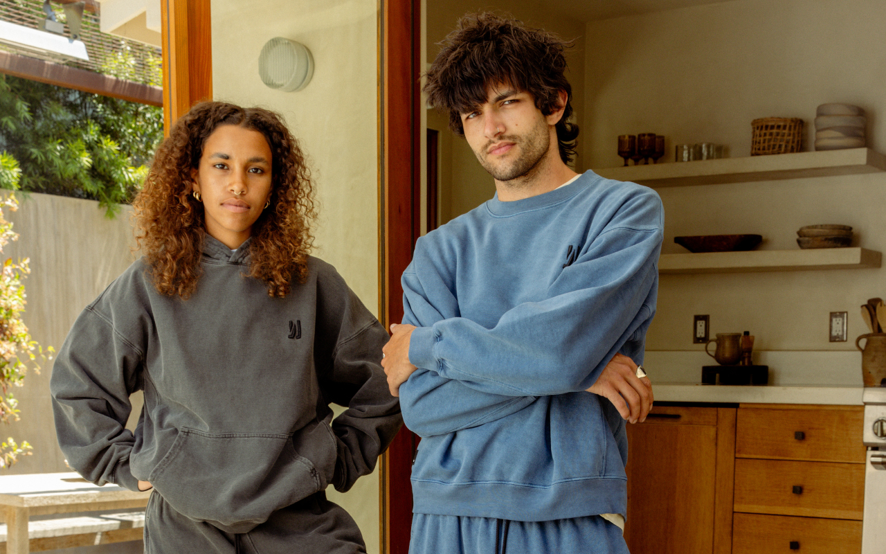 Two people standing in a kitchen, one wearing a faded black COMUNE hoodie and the other in a blue COMUNE sweatshirt.