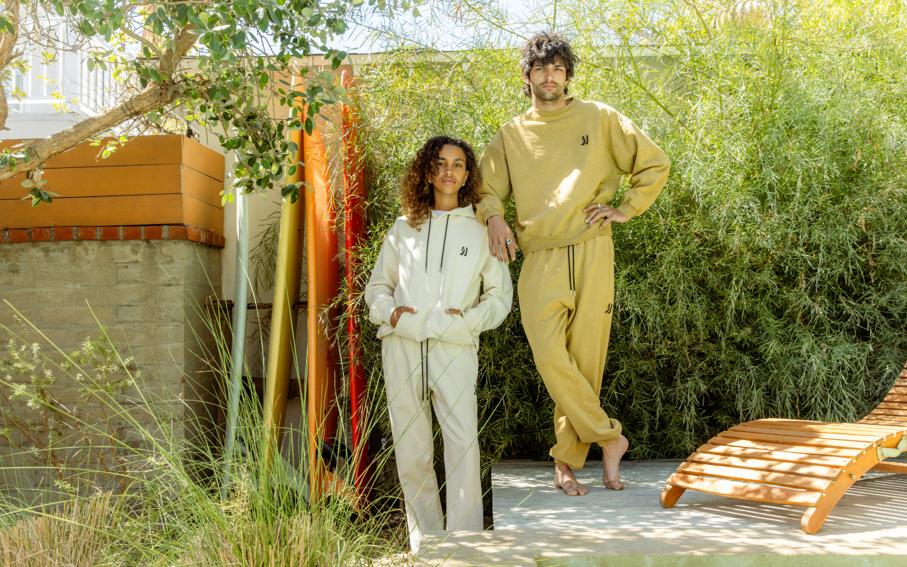 Two people in matching 'COMUNE' outfits standing outdoors with greenery in the background