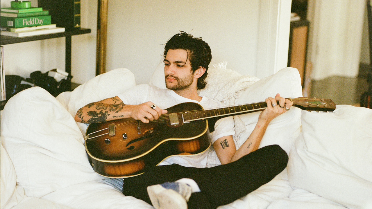 COMUNE Artist Brandon Shoop playing a guitar on a white couch in a casual setting