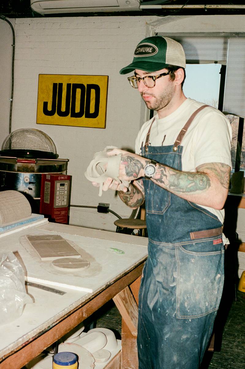 COMUNE artist Ethan Streicher wearing a 'COMUNE' cap and apron in a workshop setting working with pottery clays.
