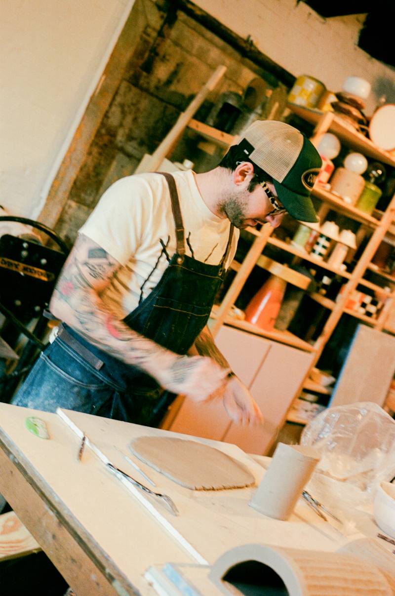 COMUNE artist Ethan Streicher wearing an apron in a workshop setting with shelves in the background