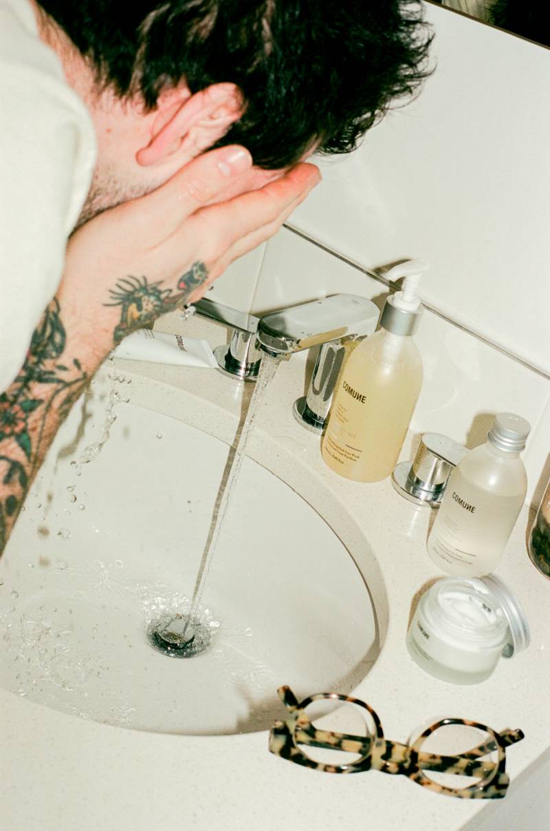 COMUNE artist Ethan  washing face with 'COMUNE' Essentials Face Cleanser in a sink, surrounded by comune products.