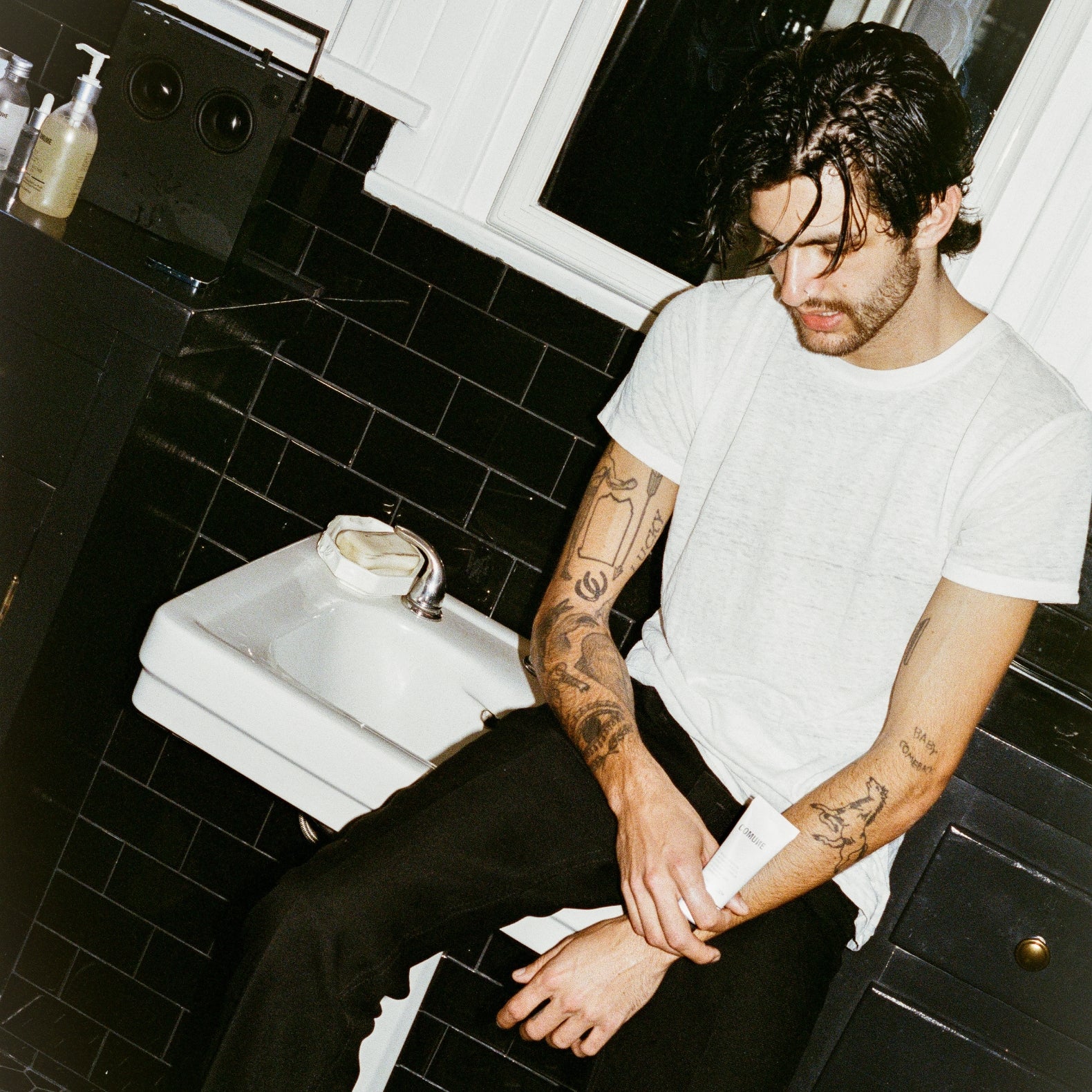 COMUNE artist Brandon sitting in a bathroom with black tiled walls and a white sink, holding 'COMUNE' Hydra Shroom Cream.