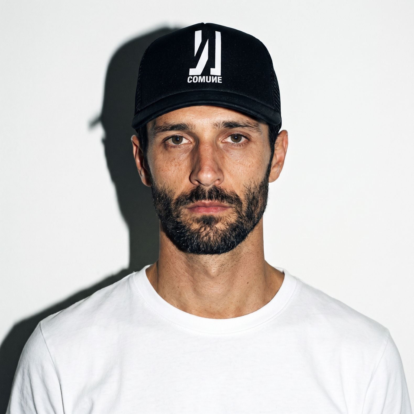 A man wearing a black COMUNE Icon active trucker hat with a white COMUNE logo and a white t-shirt, photographed against a white wall with a strong shadow.
