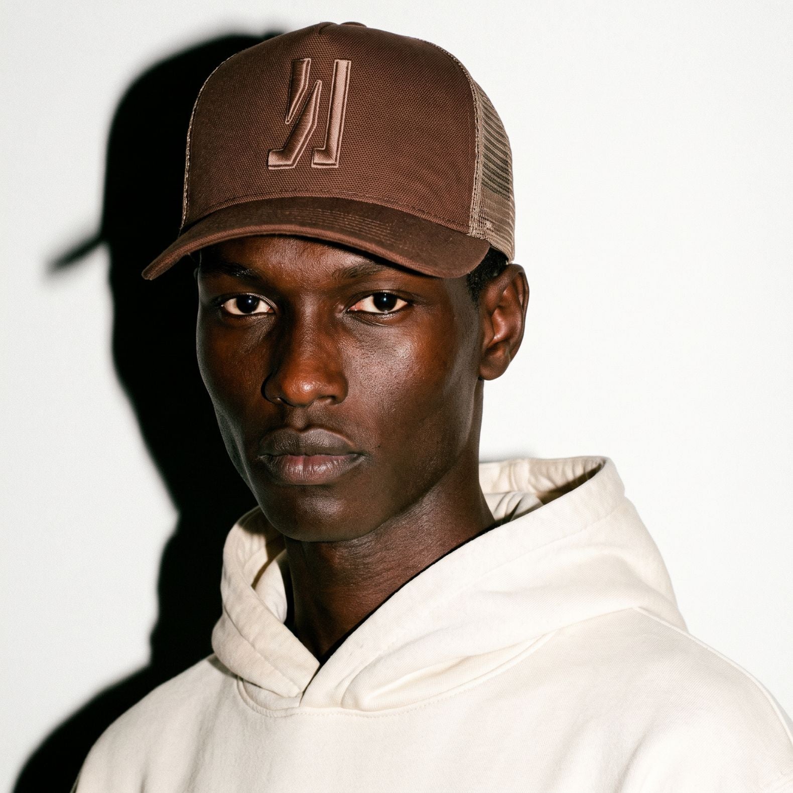 A man wearing a brown COMUNE Icon trucker cap with a tonal embroidered logo and a cream-colored hoodie, photographed against a white background with a strong shadow.