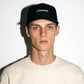 A man wearing a black COMUNE Incursive Classic baseball cap with 'Comune' embroidered in white script on the front, paired with a light beige crewneck sweatshirt, set against a plain white wall with a hard shadow.