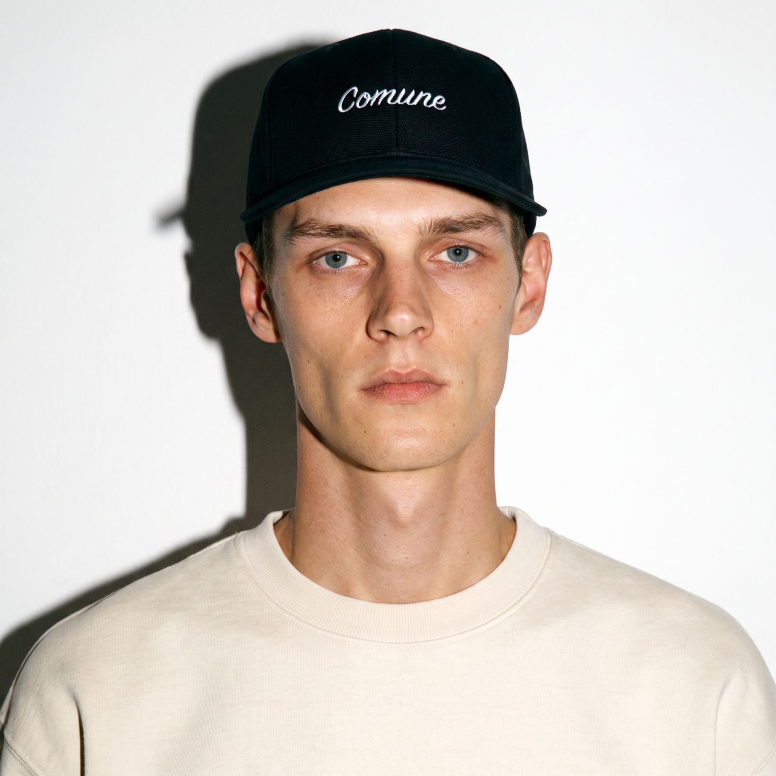 A man wearing a black COMUNE Incursive Classic baseball cap with 'Comune' embroidered in white script on the front, paired with a light beige crewneck sweatshirt, set against a plain white wall with a hard shadow.
