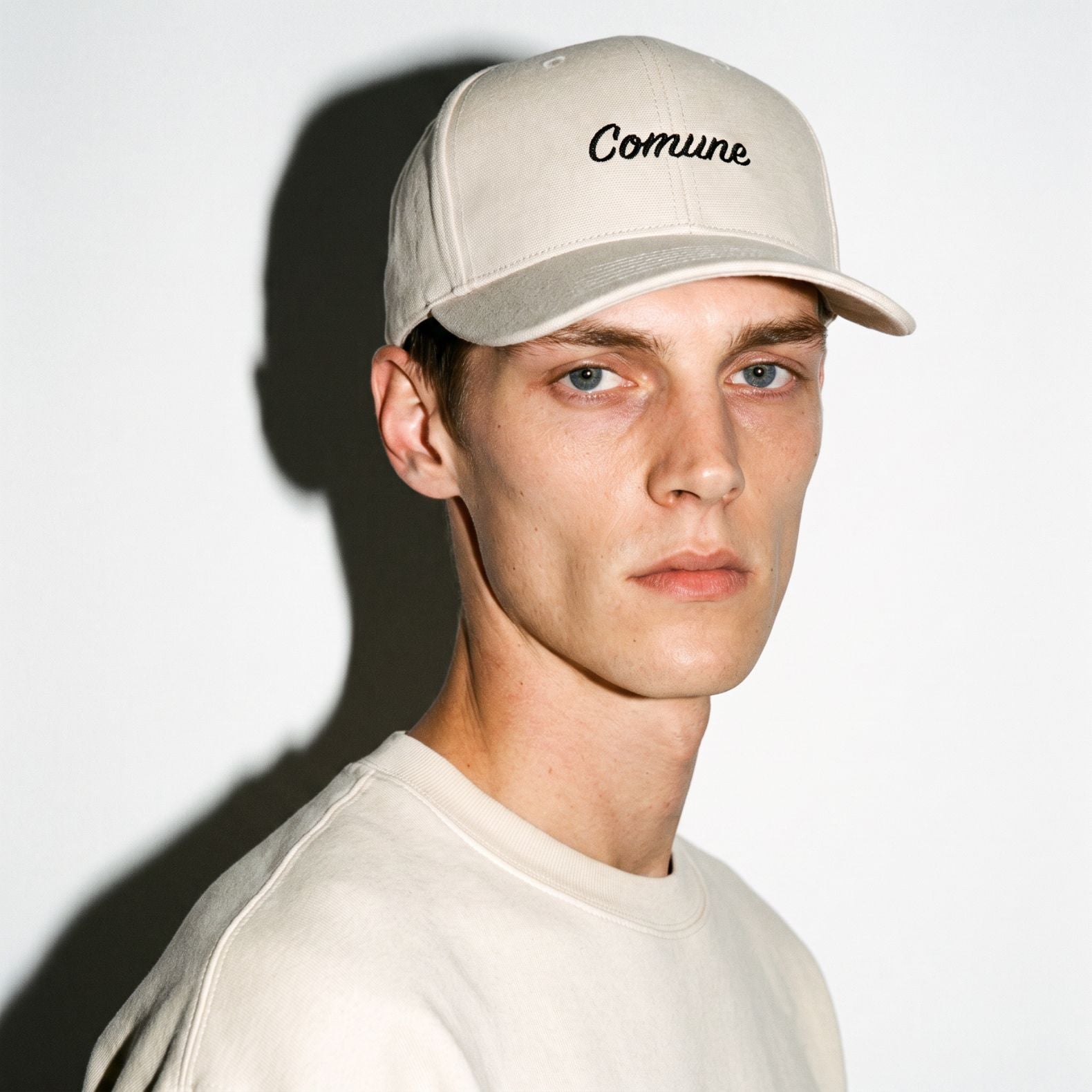 A man wearing a light beige COMUNE Incursive Classic baseball cap with the word 'Comune' embroidered in black script on the front, paired with a matching light beige crewneck sweatshirt against a white background with a strong shadow.