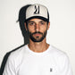 A man wearing a white crewneck t-shirt and a black and off white COMUNE Icon trucker hat with the brand name 'COMUNE' printed on the front below the Icon, set against a plain white background with a strong shadow.