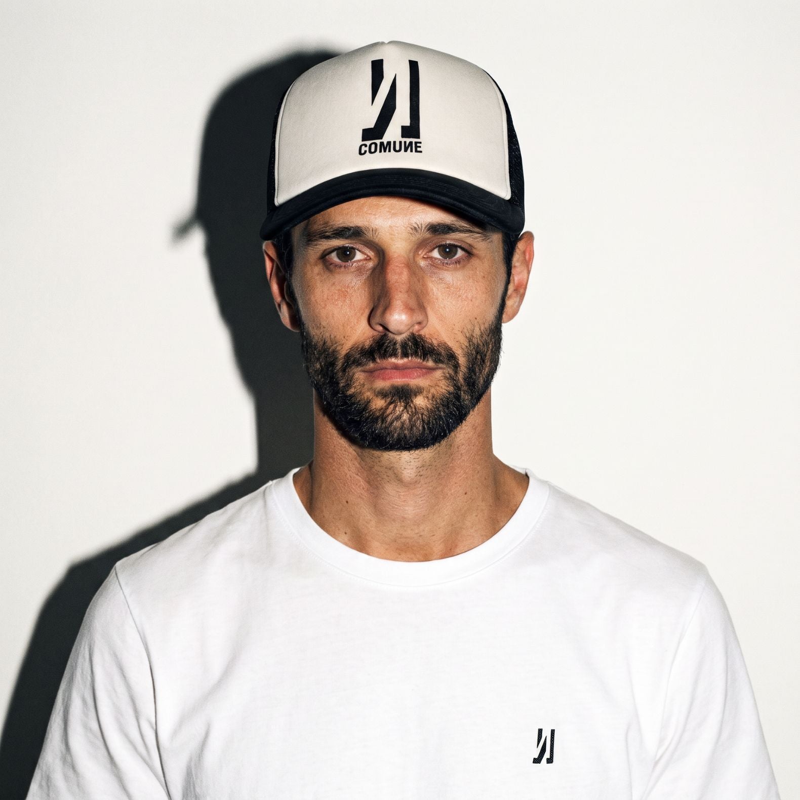 A man wearing a white crewneck t-shirt and a black and off white COMUNE Icon trucker hat with the brand name 'COMUNE' printed on the front below the Icon, set against a plain white background with a strong shadow.