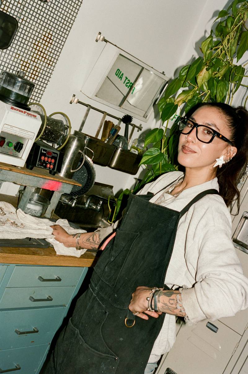 COMUNE artist Lizz Jardim wearing an apron in a work setting with plants and work tools nearby.