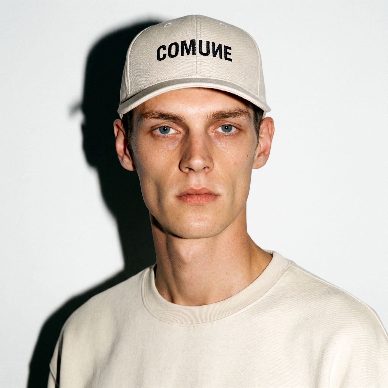 A man wearing a light beige COMUNE Logo Classic baseball cap with the word 'COMUNE' printed in black across the front, paired with a matching light beige crewneck sweatshirt against a white background with a strong shadow.