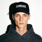 A man wearing a black COMUNE Logo trucker cap with 'COMUNE' embroidered in white on the front, paired with a dark gray washed hoodie, set against a plain white background with a strong shadow.