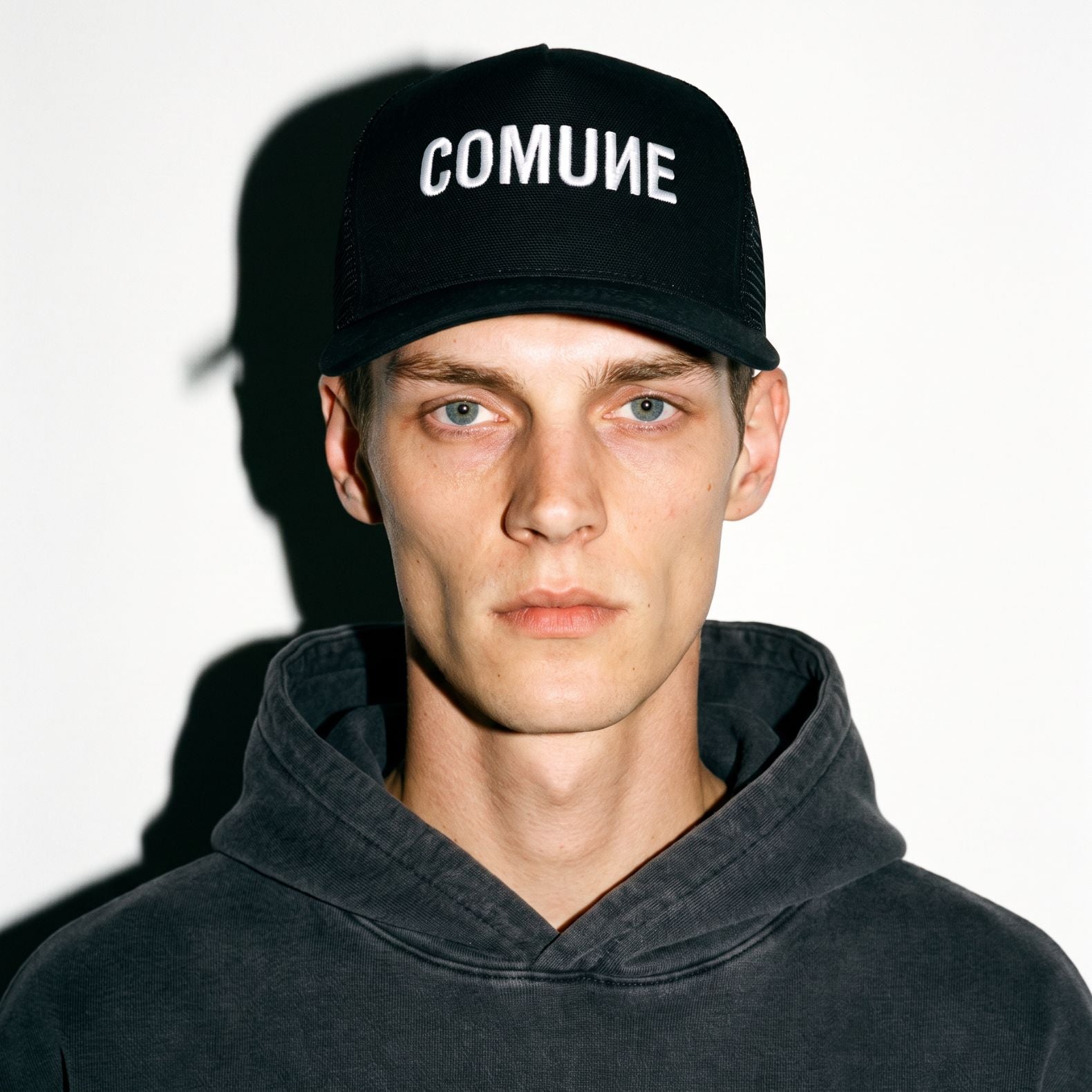 A man wearing a black COMUNE Logo trucker cap with 'COMUNE' embroidered in white on the front, paired with a dark gray washed hoodie, set against a plain white background with a strong shadow.
