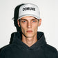 A man wearing a white COMUNE Logo trucker-style cap with the word 'COMUNE' printed in black on the front, paired with a dark charcoal hoodie, photographed against a plain white background with a strong shadow.