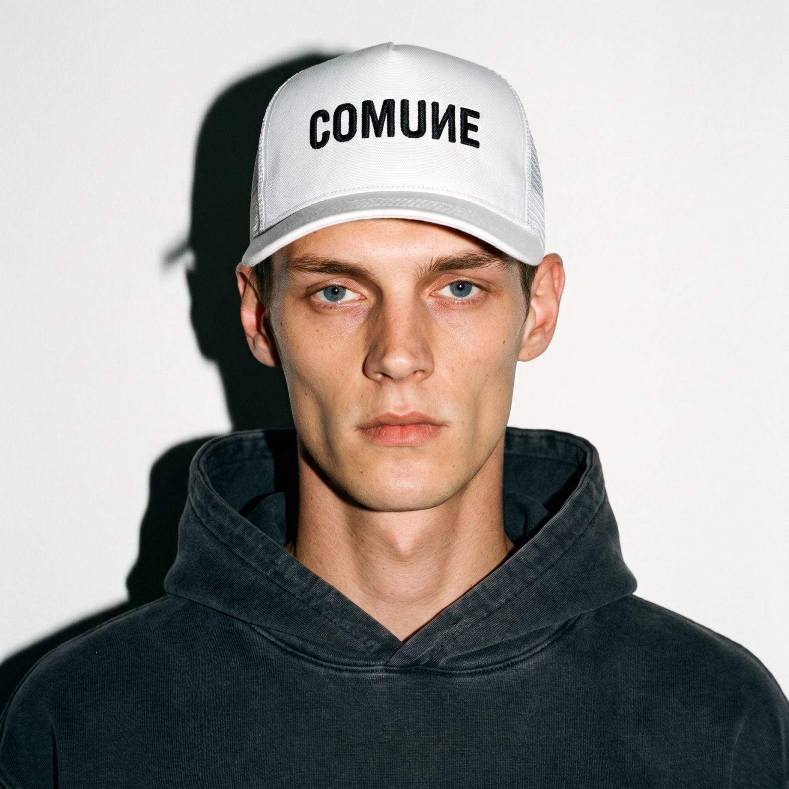 A man wearing a white COMUNE Logo trucker-style cap with the word 'COMUNE' printed in black on the front, paired with a dark charcoal hoodie, photographed against a plain white background with a strong shadow.