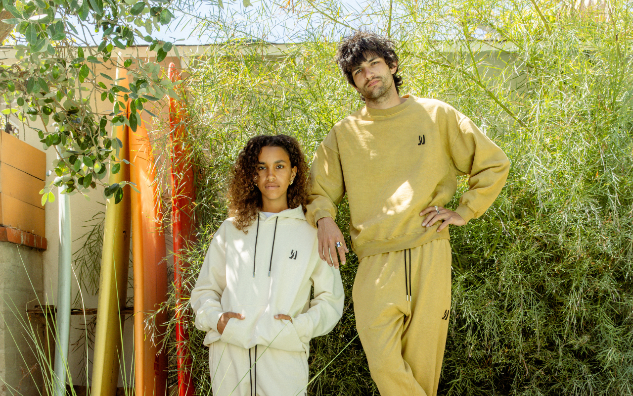 Two people wearing matching beige and white 'COMUNE' sweatshirt and sweatpants standing outdoors with greenery in the background.