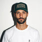 A man wearing a hunter green COMUNE Mission trucker hat with the text 'SOMETHING BETTER CHANGE' in beige, paired with a white t-shirt, photographed against a plain white wall with a strong shadow.