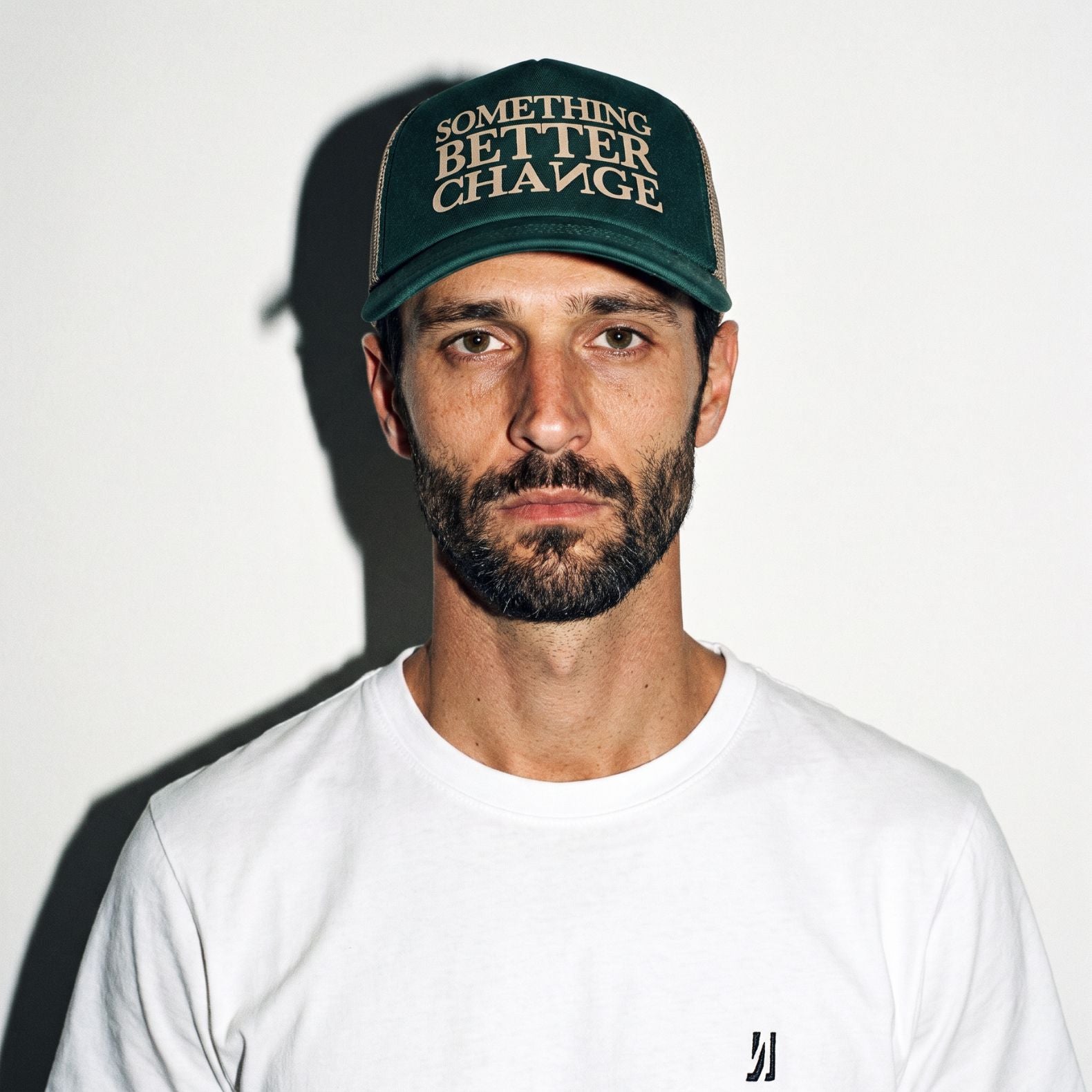 A man wearing a hunter green COMUNE Mission trucker hat with the text 'SOMETHING BETTER CHANGE' in beige, paired with a white t-shirt, photographed against a plain white wall with a strong shadow.