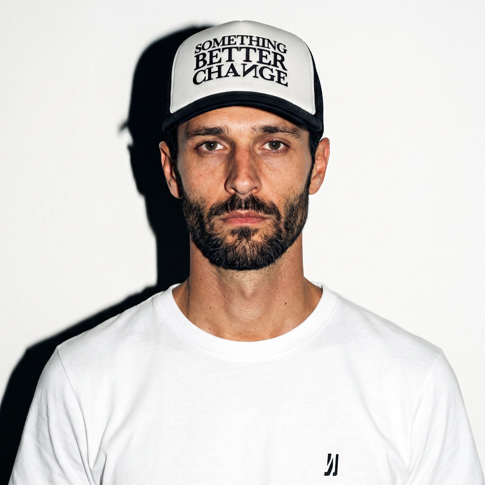 A man wearing an off white COMUNE Mission trucker hat with black lettering that reads 'SOMETHING BETTER CHANGE' and a white t-shirt, photographed against a white background with a strong shadow.