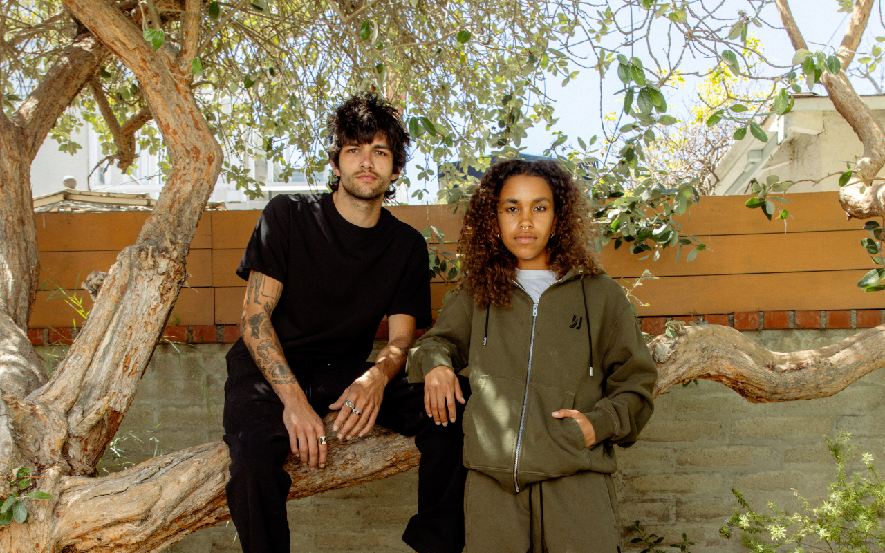 Two people sitting on a tree branch with a natural background wearing COMUNE clothing.