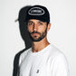 A man wearing a black COMUNE Patch trucker hat with 'COMUNE' in white text on the front, paired with a white t-shirt, set against a plain white wall with a strong shadow.