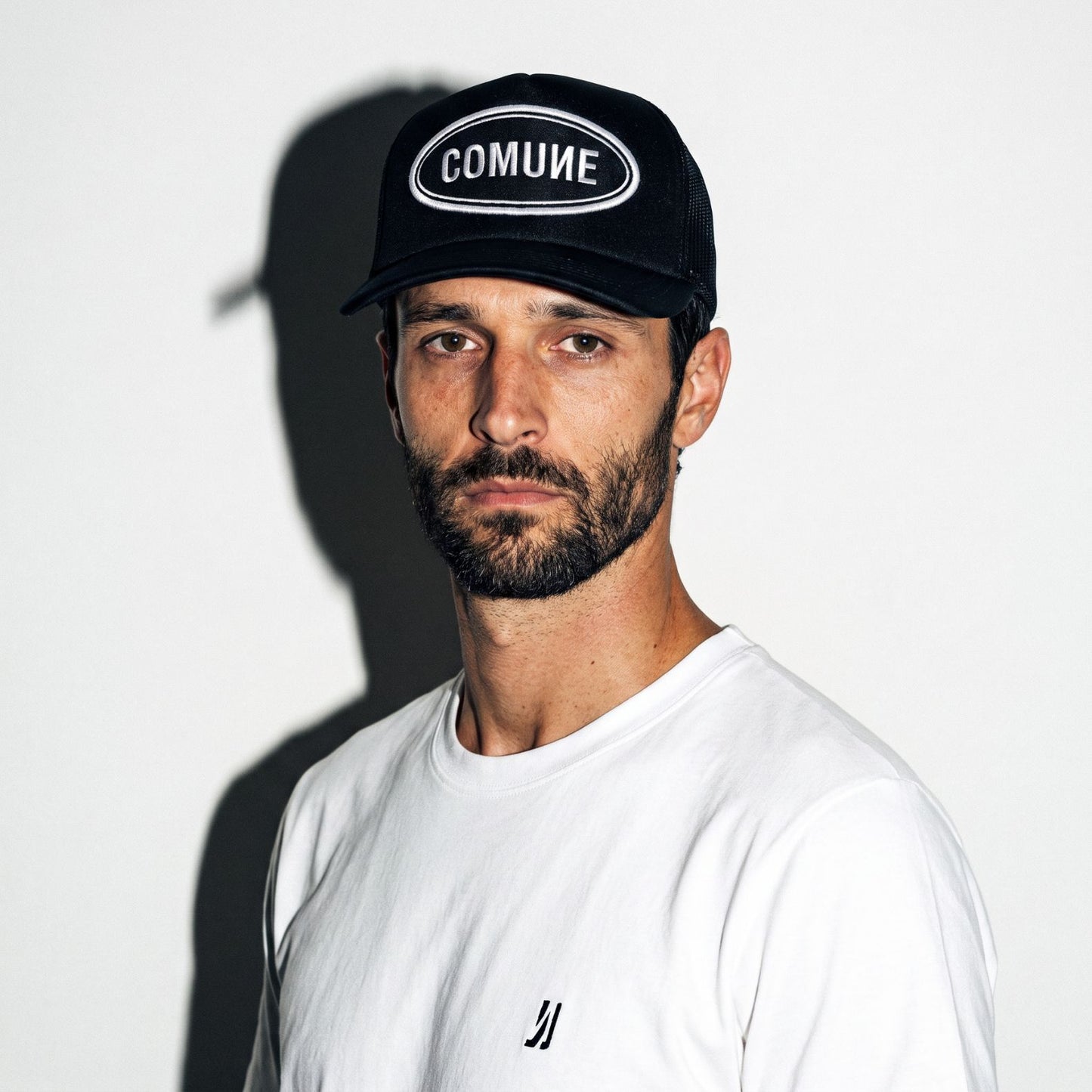 A man wearing a black COMUNE Patch trucker hat with 'COMUNE' in white text on the front, paired with a white t-shirt, set against a plain white wall with a strong shadow.