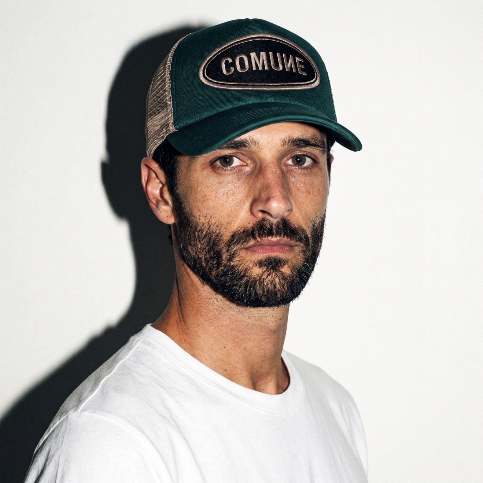 A man wearing a hunter green COMUNE Patch trucker-style baseball cap with a black and beige patch reading 'COMUNE' and a white t-shirt, photographed against a plain white wall with a strong shadow.