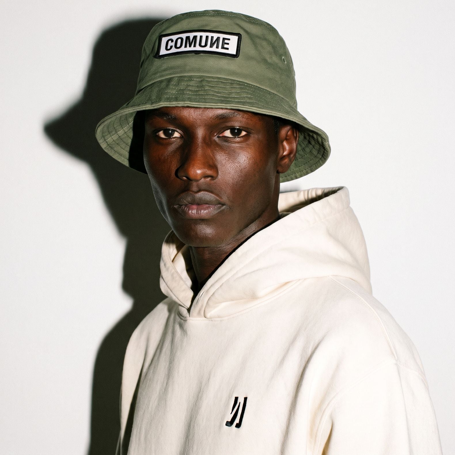 A man wearing a army green COMUNE Patch bucket hat with a white 'COMUNE' patch and a cream-colored hoodie, photographed against a white wall with a strong shadow.