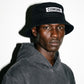 A man wearing a black COMUNE Patch bucket hat with a white rectangular patch that reads 'COMUNE' in black letters, paired with a faded black hoodie, photographed against a white background with a strong shadow.