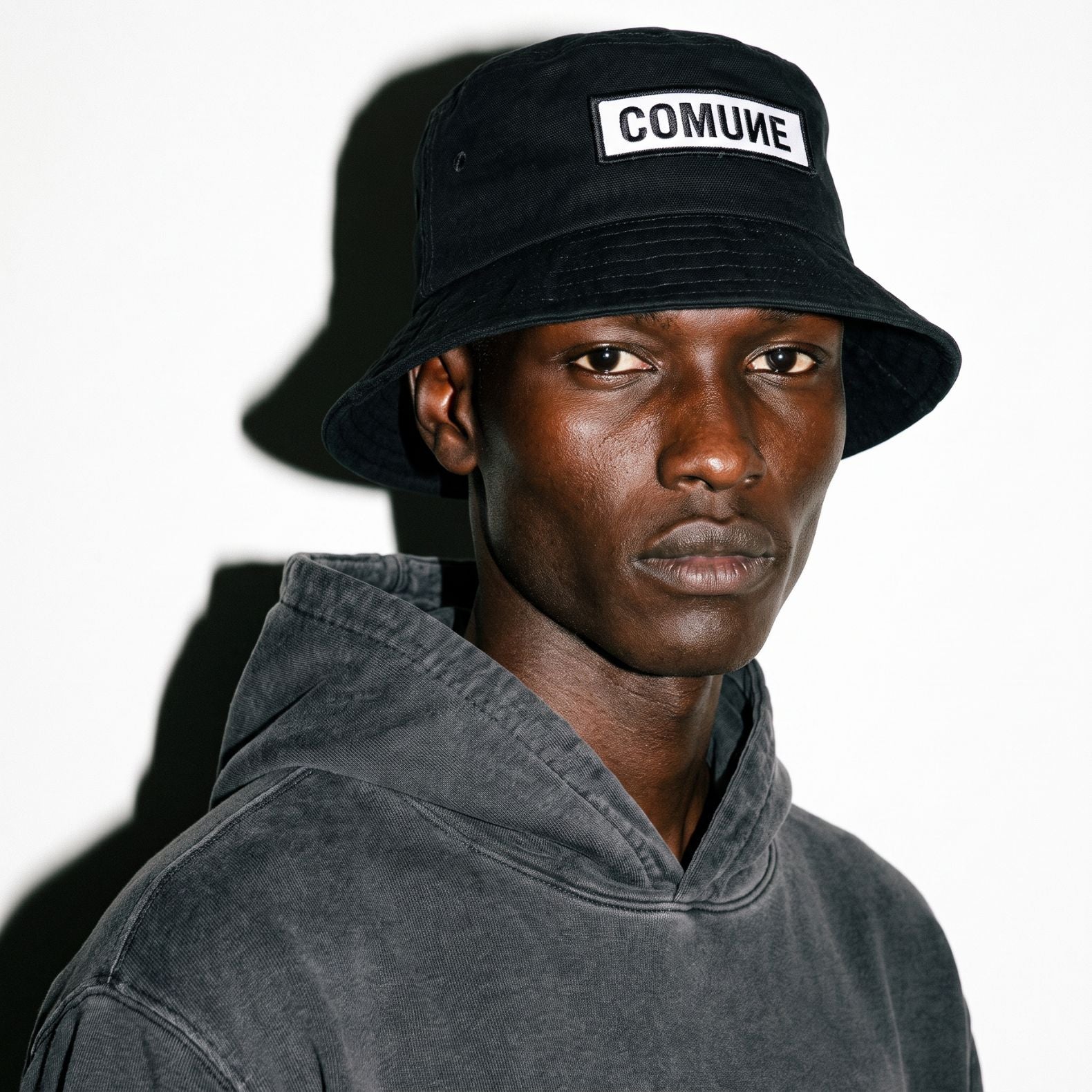 A man wearing a black COMUNE Patch bucket hat with a white rectangular patch that reads 'COMUNE' in black letters, paired with a faded black hoodie, photographed against a white background with a strong shadow.