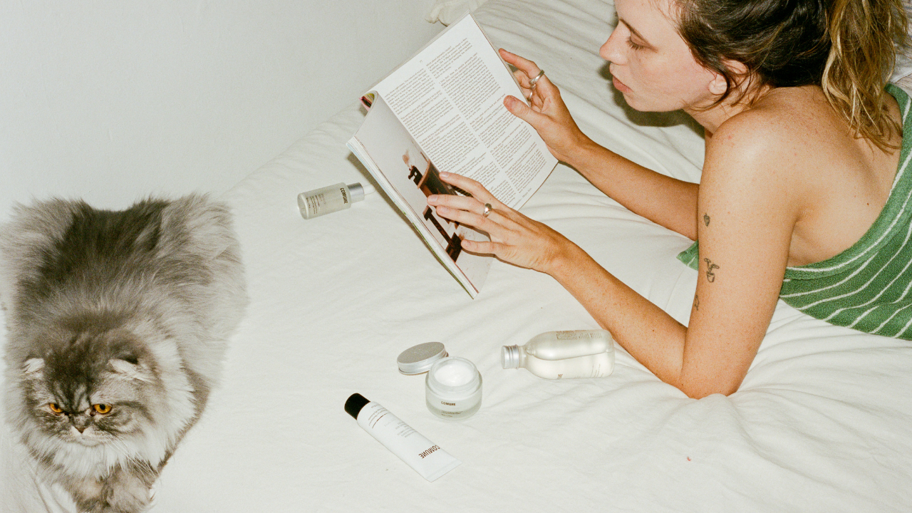 COMUNE model reading a book on a bed with a cat and various 'COMUNE' skincare products around