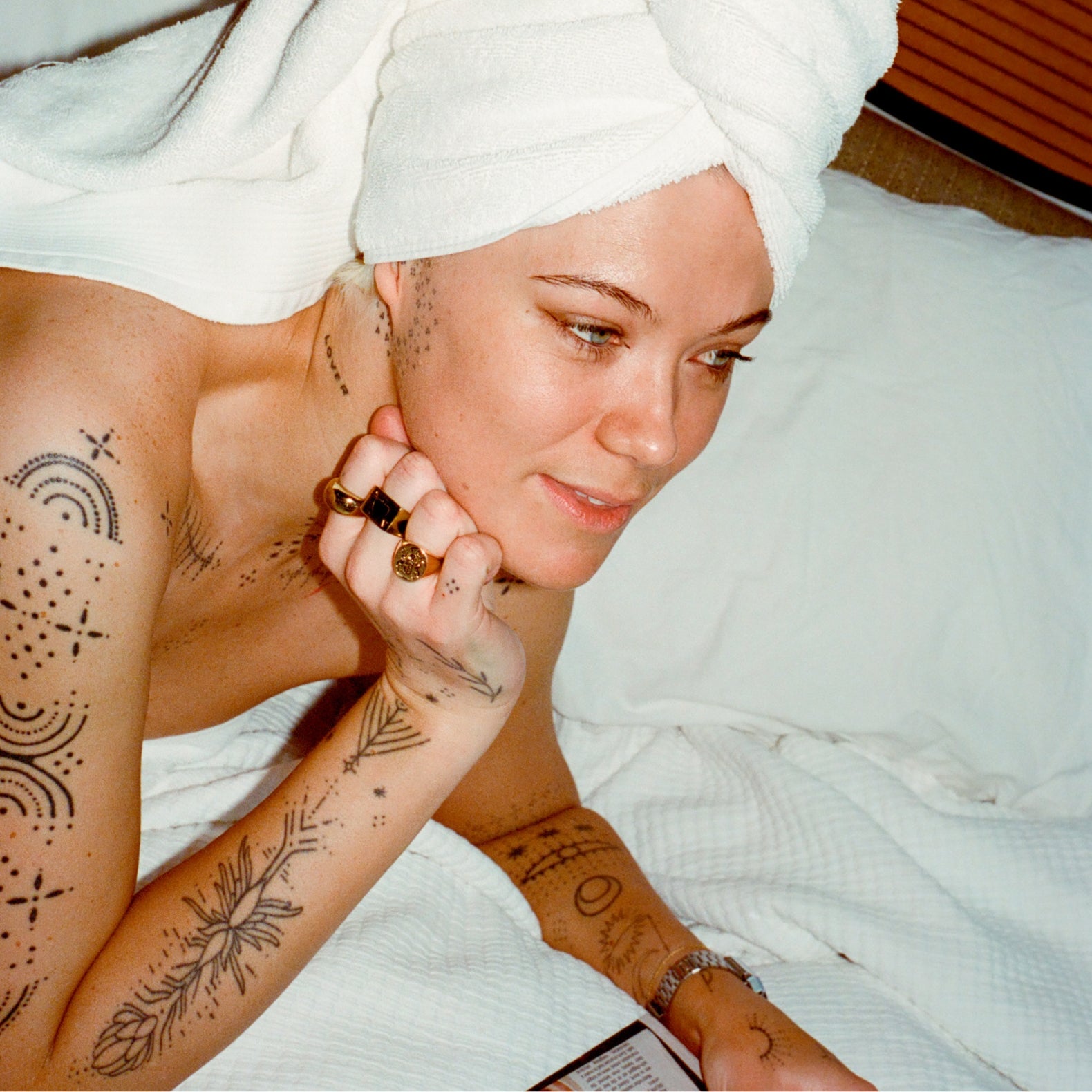 Woman with tattoos on her arms and chest, wearing a white towel, sitting on a bed.