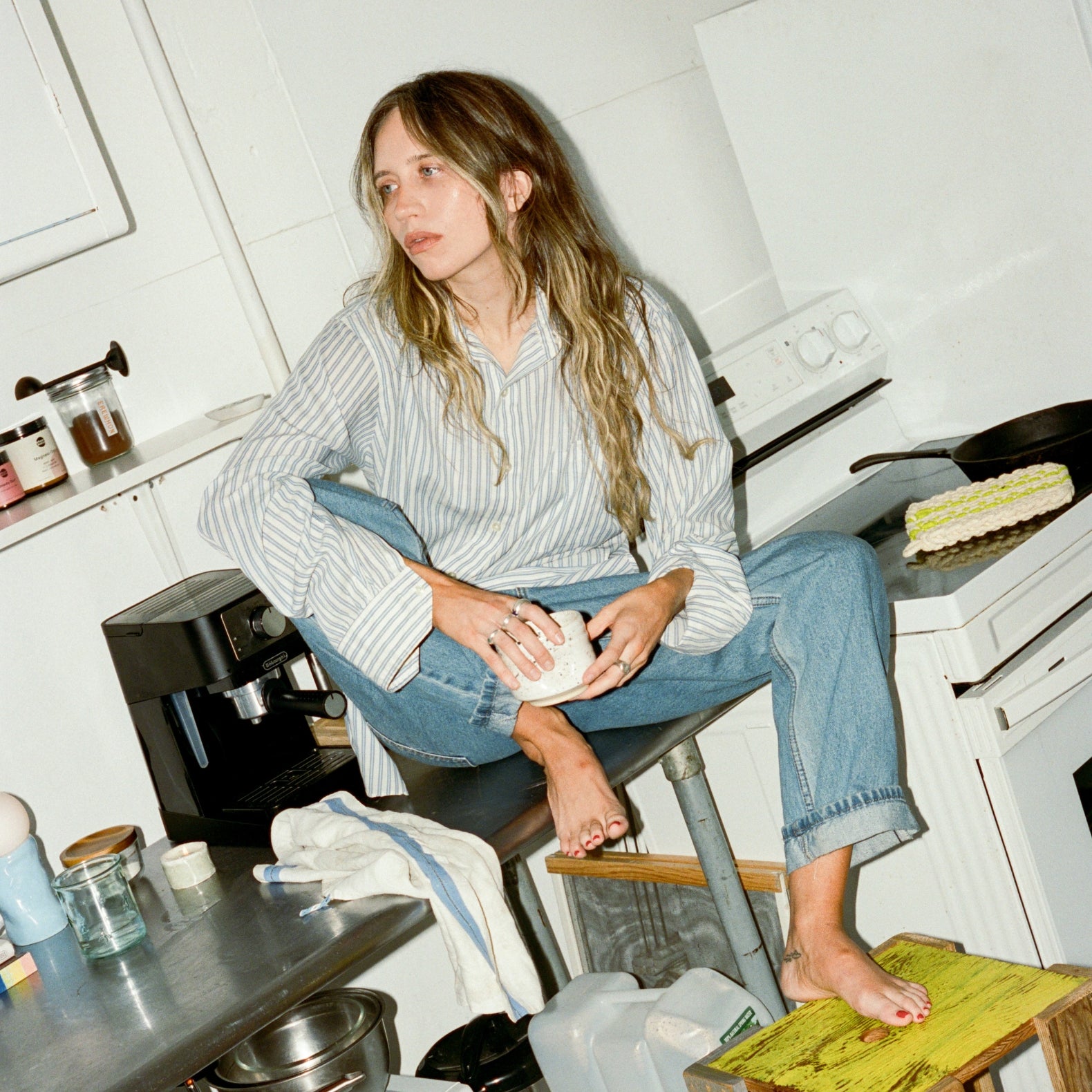 Woman sitting on a kitchen counter holding a white object, surrounded by kitchen appliances and items.
