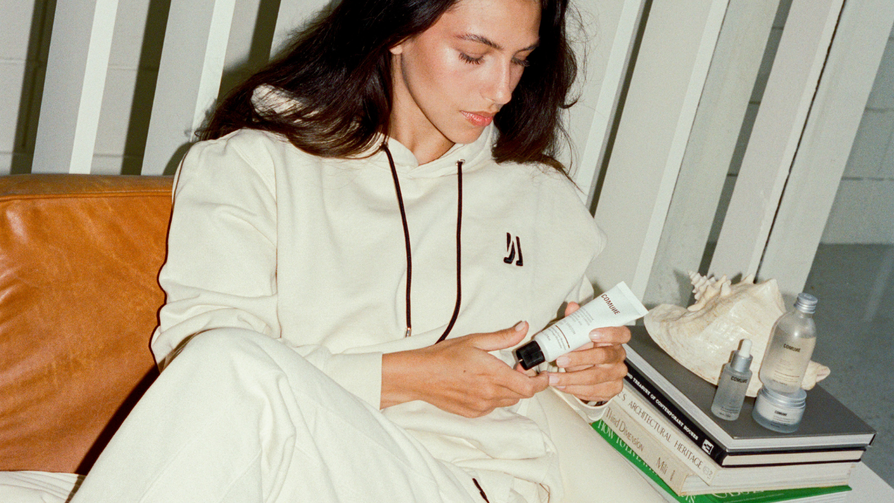 COMUNE model wearing COMUNE clothing sitting on a couch holding a Hydra Shroom Cream product, with books and bottles on a table beside her.