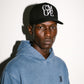 A man wearing a black COMUNE Stack trucker hat with a white graphic logo and a washed blue hooded sweatshirt, photographed against a plain white wall with a strong shadow.
