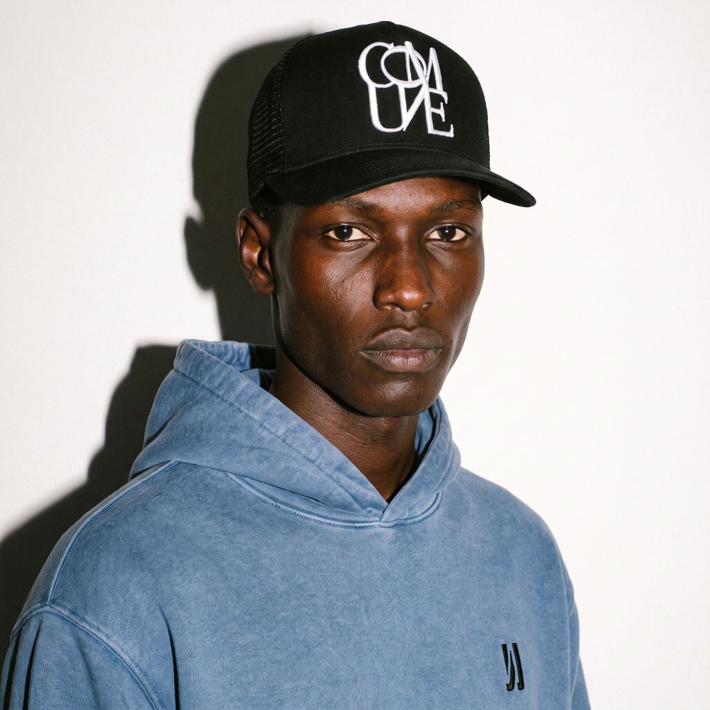 A man wearing a black COMUNE Stack trucker hat with a white graphic logo and a washed blue hooded sweatshirt, photographed against a plain white wall with a strong shadow.