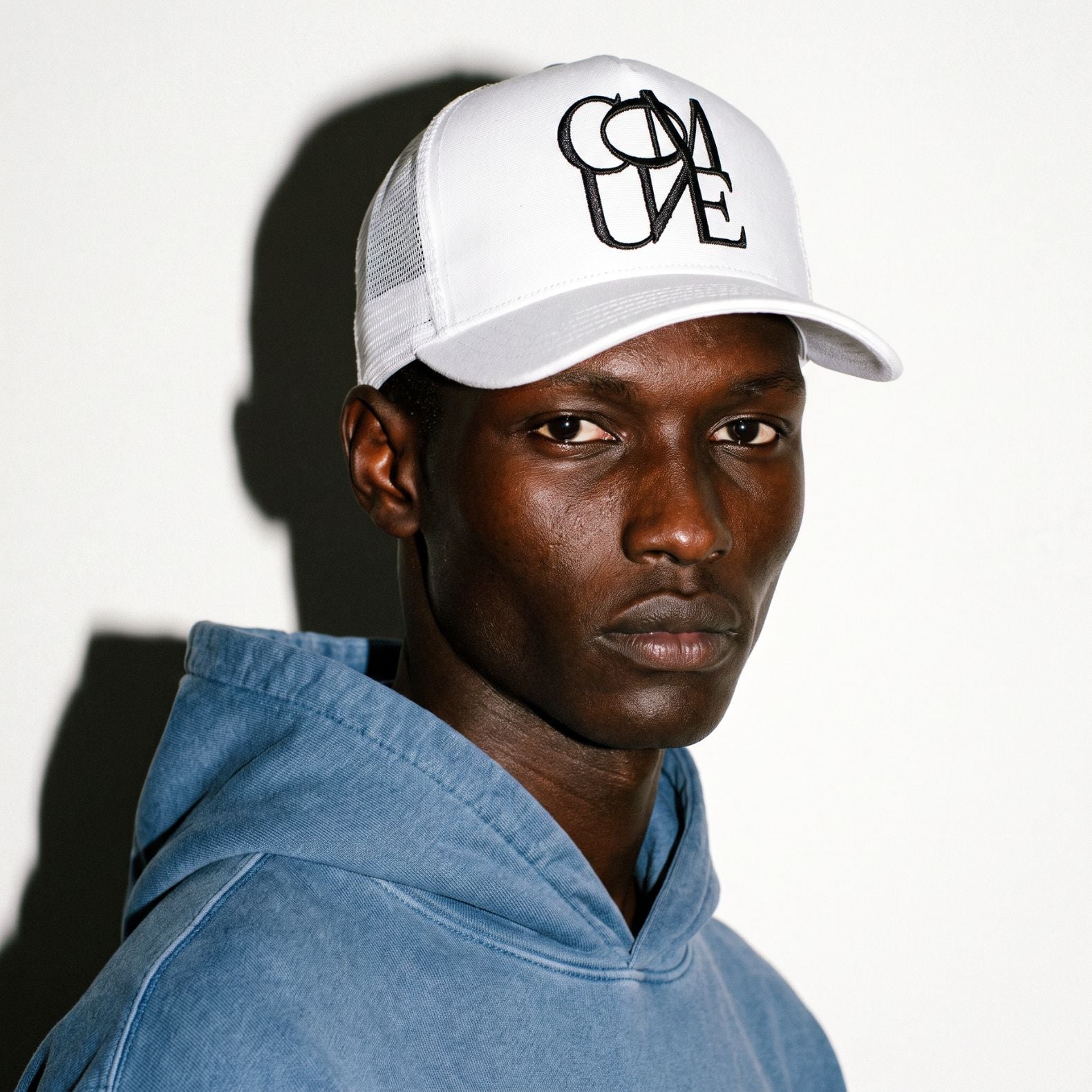 A man wearing a white COMUNE Stack trucker-style cap with a black stylized 'COMUNE' graphic on the front, paired with a blue hooded sweatshirt, photographed against a plain white wall with a strong shadow.