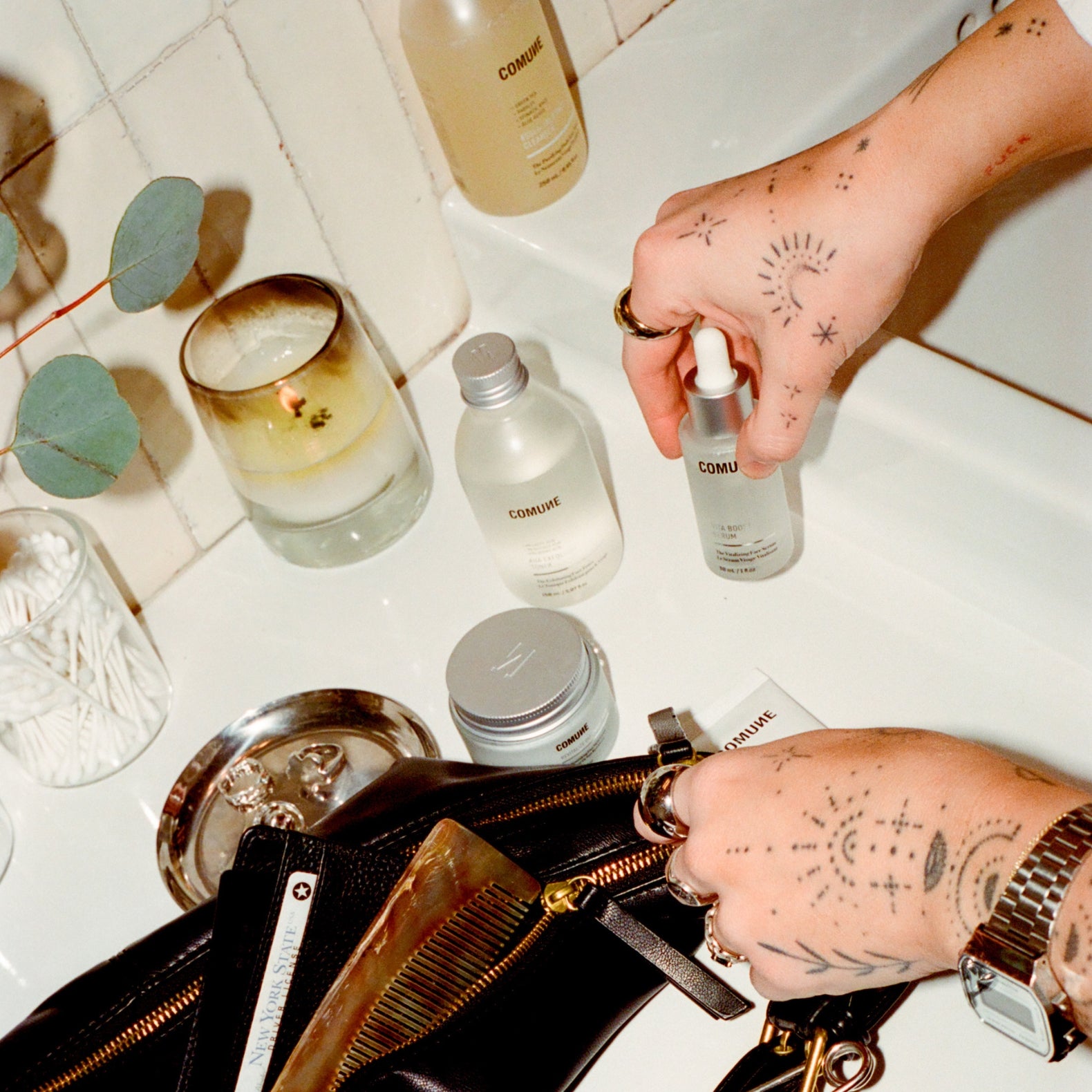 Person holding a bottle of 'COMUNE' skincare product on a bathroom counter with various items.