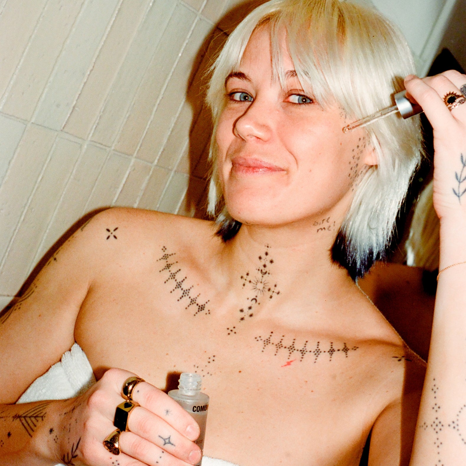 Comune model with tattoos applying comune's vita boost serumt to her face in a bathroom setting