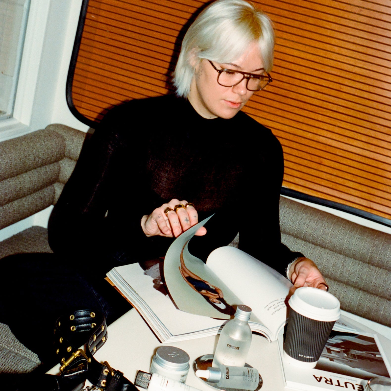 Comune model with glasses reading a magazine in a living room with 'COMUNE' essentials skincare products on the table.