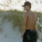 Man walking on a beach with a surfboard, wearing a COMUNE cap and black shorts.