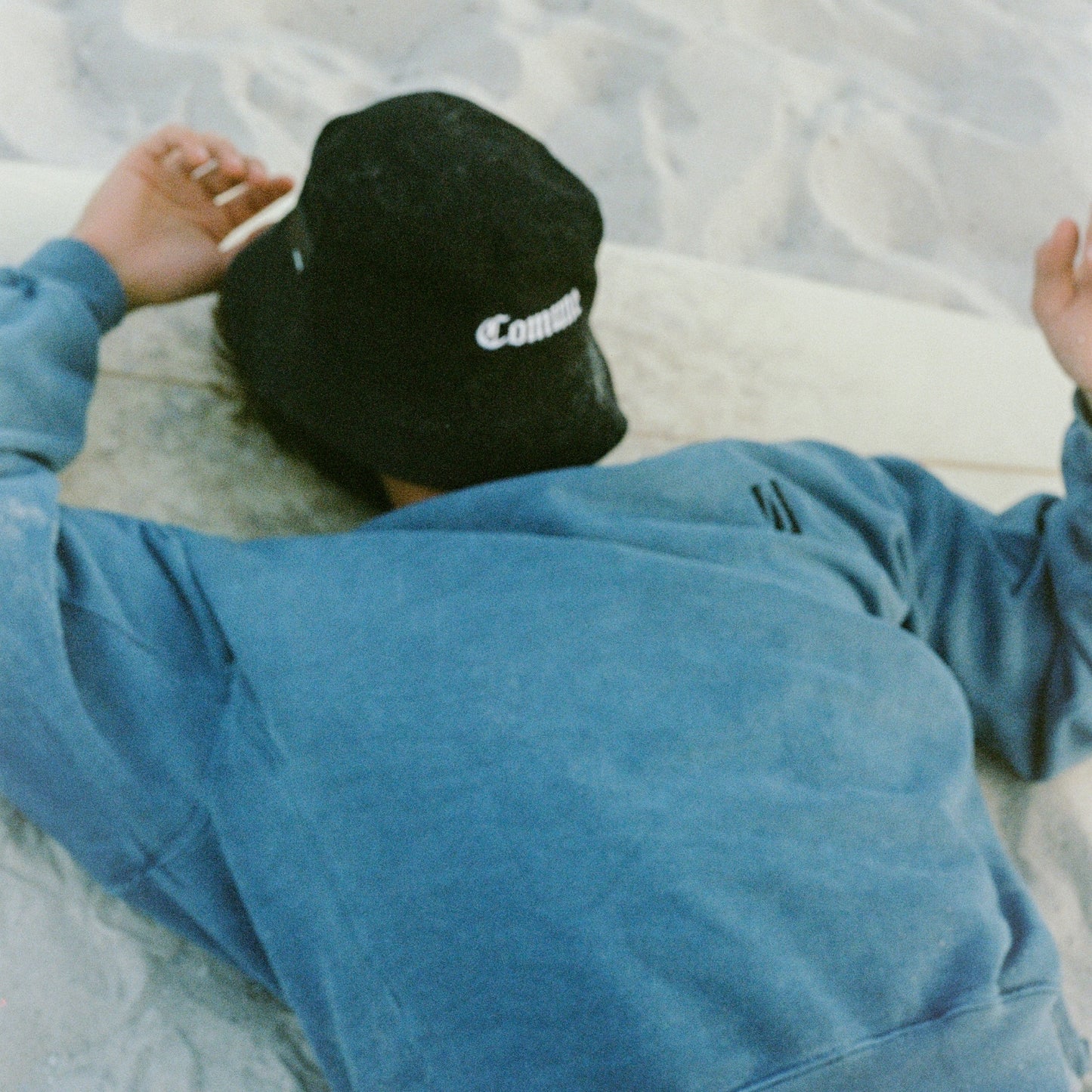 Person wearing a COMUNE black bucket hat with 'COMUNE' logo and COMUNE Duke blue sweatshirt lying at the beach..