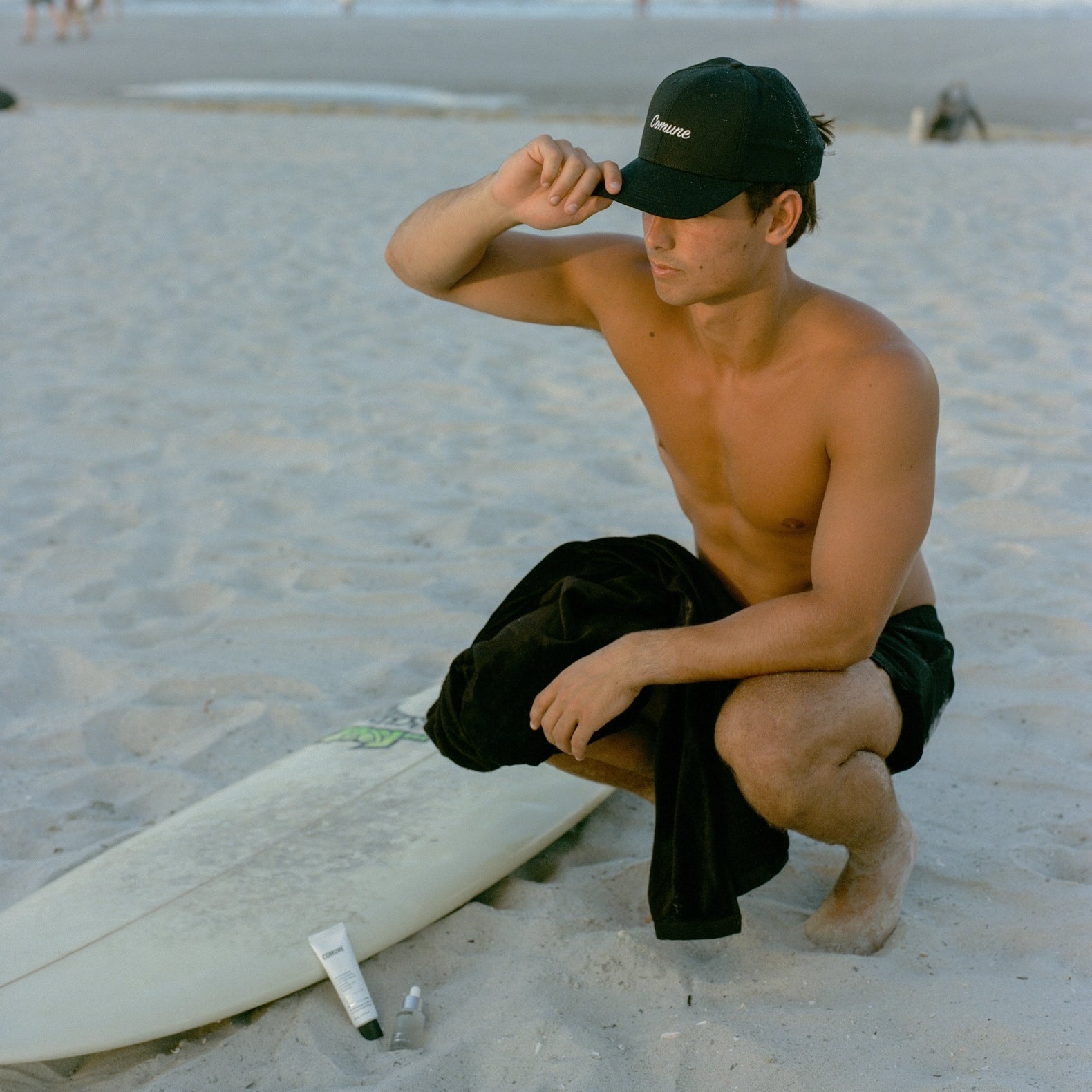 Man sitting on the sand with a surfboard, wearing a black COMUNE classic cap and shorts.