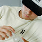 Person wearing a bone color COMUNE Duke Crew heavyweight cotton sweatshirt and the Icon Active trucker hat, holding a bottle of COMUNE AHA Exfoliant Toner.