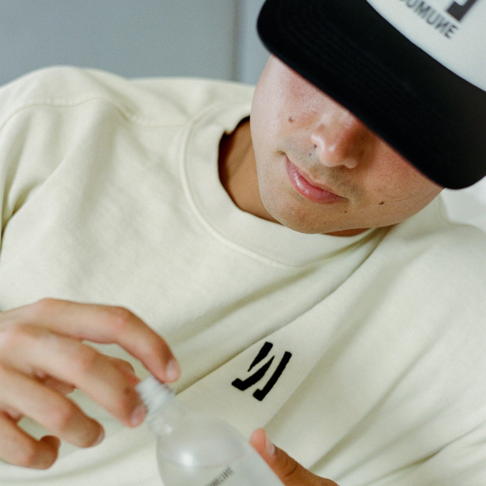 Person wearing a bone color COMUNE Duke Crew heavyweight cotton sweatshirt and the Icon Active trucker hat, holding a bottle of COMUNE AHA Exfoliant Toner.
