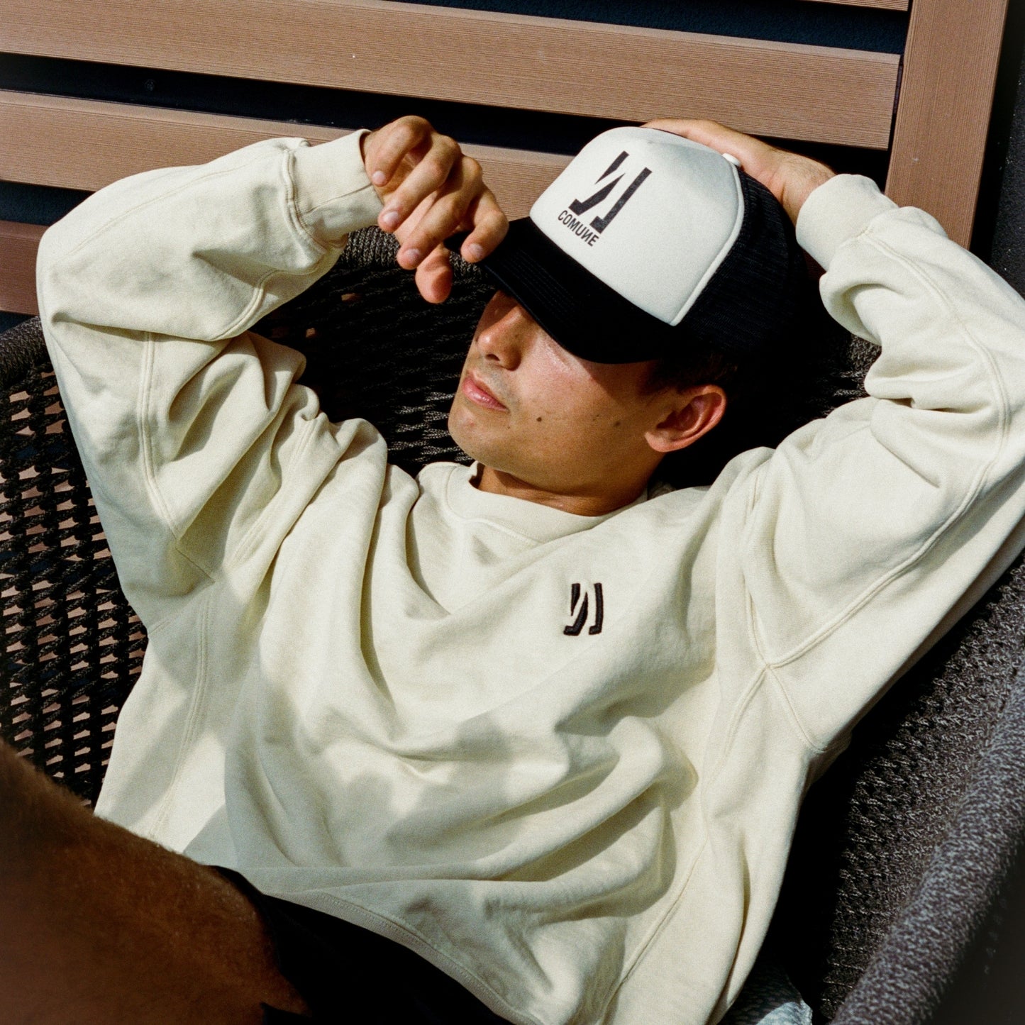 Surfer wearing a COMUNE Duke sweatshirt and Comune Icon Trucker cap, sitting on a wicker chair.