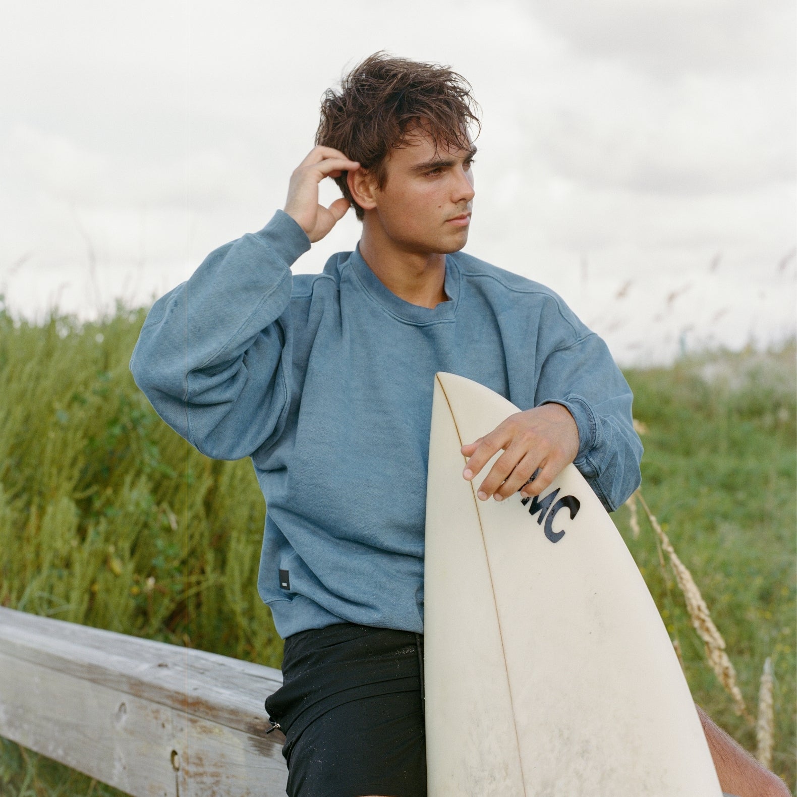 Man holding a surfboard wearing COMUNE Duke Crew sweatshirt in marine color in a grassy field