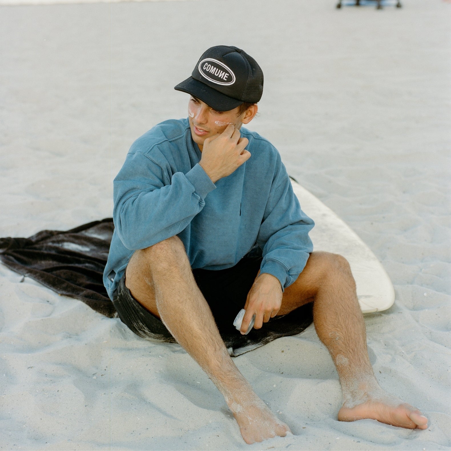 Surfer sitting on a sandy beach with a surfboard, wearing a COMUNE Duke sweatshirt and COMUNE Logo Trucker black cap, applying COMUNE face cream.